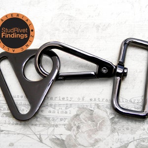 May include: A black metal triangle swivel trigger snap hook with a rectangular D-ring. The hook is attached to the D-ring. The metal is shiny and has a black finish. The text "StudRivet Findings" is printed on a circle in the background.