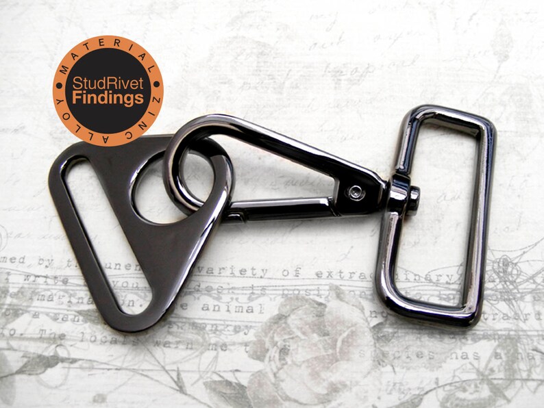 May include: A black metal triangle swivel trigger snap hook with a rectangular D-ring. The hook is attached to the D-ring. The metal is shiny and has a black finish. The text "StudRivet Findings" is printed on a circle in the background.