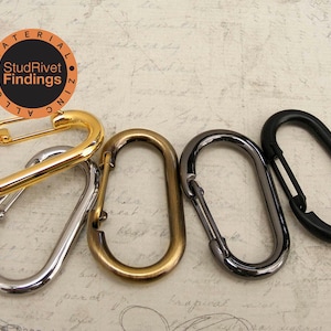 May include: Five oval-shaped metal clasps in different finishes: gold, silver, antique brass, black, and gunmetal. The clasps are all the same size and shape, and they are arranged in a semi-circle on a textured background. The text "StudRivet Findings" is visible in a circle on the top left of the image.