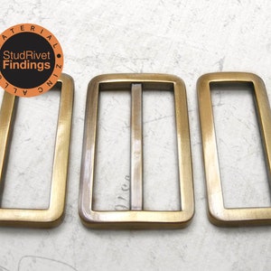 May include: Three antique brass-coloured rectangular belt buckles and loops. The centre buckle has a bar. The image includes a circular logo with the text "MATERIAL ALLOY ZINC FINDS StudRivet".