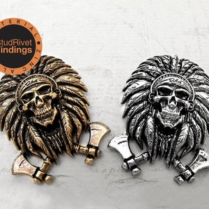 May include: Two metal skull pendants with Native American headdresses and crossed axes. The pendant on the left is gold, and the pendant on the right is silver.