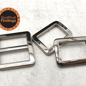 May include: Three silver-colored, square metal buckles. One buckle has a center bar. The buckles are shiny and reflective. The image includes a circular logo with the text "StudRivet Findings".