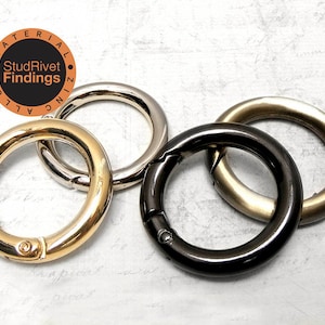May include: Four metal round spring gate rings in gold, silver, black, and brass finishes. The rings are of varying sizes and have a spring-loaded clasp mechanism. The StudRivet Findings logo is in the upper left corner.