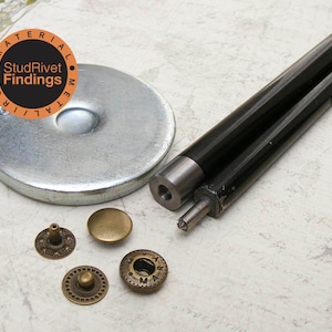 Peut inclure: Un ensemble de rivets métalliques et un outil pour les poser. L'outil est noir et a une poignée argentée. Les rivets sont dorés et ont un design décoratif. L'ensemble est étiqueté "StudRivets Findings Material Metal Iron".