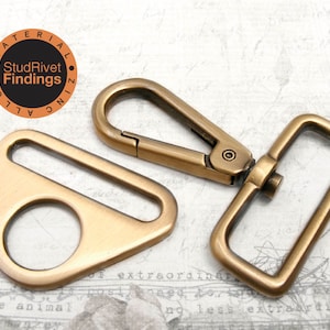 May include: A set of two antique gold metal hardware pieces for bags or other crafts. The set includes a triangular swivel hook and a rectangular D-ring.