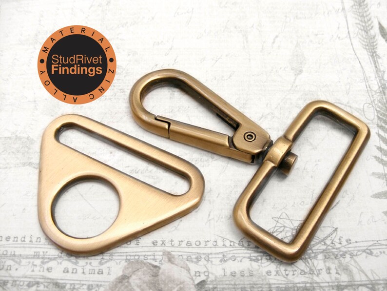 May include: A set of two antique gold metal hardware pieces for bags or other crafts. The set includes a triangular swivel hook and a rectangular D-ring.