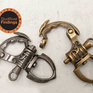 May include: Two decorative metal swivel clasps in antique bronze and silver. The clasps have detailed patterns and are labelled "StudRivet Findings". These clasps are likely for jewellery making or leathercraft projects.