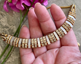 May include: A gold-toned necklace with a curved design, featuring alternating sections of clear crystals and rectangular mother-of-pearl inlays. The necklace is held in a hand, with pink flowers in the background.
