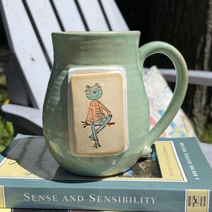 Puede incluir: Una taza de cerámica verde claro con una ilustración de una rana en un parche rectangular. La rana lleva una bufanda y sostiene una taza de café. La taza está sobre una pila de libros, incluyendo "Sense and Sensibility".