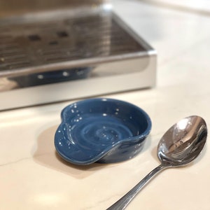 May include: A blue ceramic spoon rest with a swirl design. The spoon rest is sitting on a white countertop next to a silver spoon.