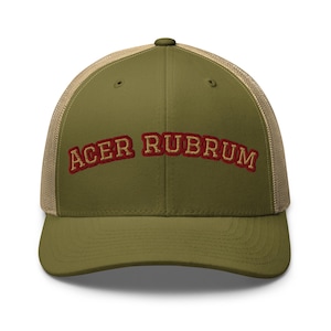 Könnte beinhalten: Eine olivgrüne Baseballkappe mit beigem Netzrücken und strukturiertem Vorderteil. Der Text ACER RUBRUM ist vorne in goldener Schrift mit dunkelroter Umrandung aufgestickt, mit einem geschwungenen Design und einem Knopf an der Oberseite.
