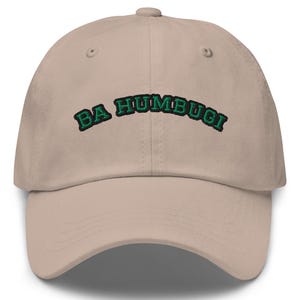 Peut inclure: Une casquette de baseball beige avec le texte brodé BA HUMBUG! en vert foncé avec un contour noir. La casquette présente une calotte structurée, une visière incurvée et un bouton sur le dessus, avec des détails de couture visibles.