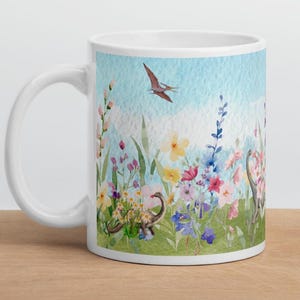 May include: White ceramic mug featuring a watercolor design of a field of colorful flowers, dinosaurs, and a flying pterodactyl against a blue sky. The mug has a white handle and a glossy finish.