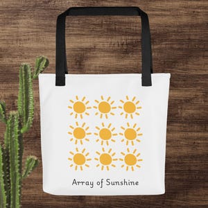 Könnte beinhalten: Weiße Einkaufstasche mit schwarzen Henkeln und einem Design aus 12 gelben Sonnen in einem Rastermuster. Der Text "Array of Sunshine" ist unter den Sonnen gedruckt.