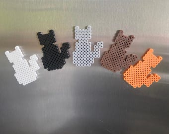 May include: Four small cat-shaped bead ornaments in white, black, brown, and orange. The ornaments are arranged in a row on a silver surface.
