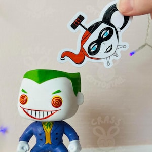 May include: A white vinyl sticker featuring a cartoon illustration of Harley Quinn, a DC Comics character. Harley Quinn is depicted with red and black hair, a black mask, and a red and white jester's hat. The sticker is being held by a hand over a figurine of the Joker, a DC Comics character.
