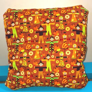 May include: A square throw pillow featuring a repeating pattern of scarecrows, orange pumpkins, and yellow sunflowers on a textured brown background. The scarecrows wear various outfits including hats, plaid shirts, and dresses in shades of yellow, green, and orange.