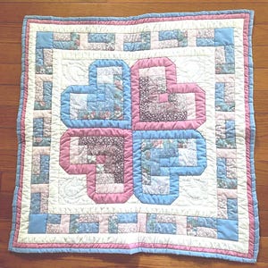 May include: A square patchwork quilt featuring a central design of four interlocking heart shapes constructed from log cabin blocks in shades of light blue, dusty pink, and floral patterns, surrounded by a cream border and a blue outer edge.