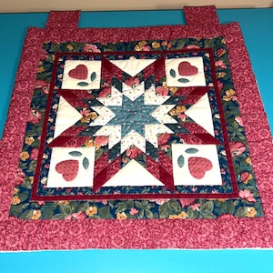 May include: A square quilted wall hanging featuring a central star pattern in shades of blue, cream, and deep red. The design includes four corner squares with red heart motifs, surrounded by a floral border and two fabric tabs at the top edge.