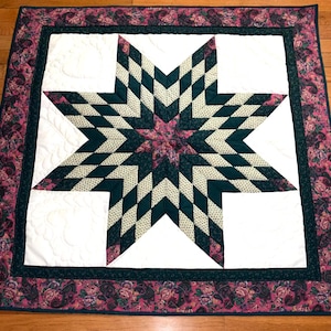 May include: A square quilt featuring a central eight-pointed star pattern composed of dark teal and cream-colored fabric diamonds. The star is set against a white background with quilted heart motifs, framed by a dark teal border and a floral paisley border.