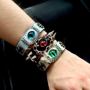 May include: Three leather bracelets with eye designs. One is light blue with a blue eye, another is black with a red eye, and the third is light green with a green eye. Each bracelet has metal accents and chains.