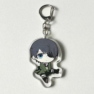 May include: A clear acrylic keychain featuring a cartoon character with black hair, a green outfit, and an eyepatch. The character is sitting with one leg crossed over the other.