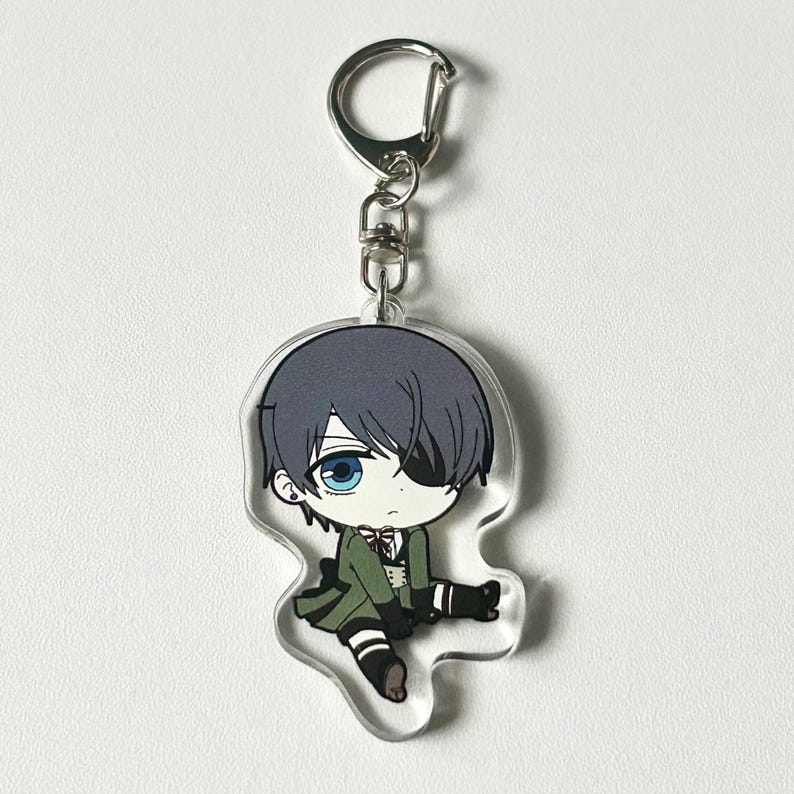 May include: A clear acrylic keychain featuring a cartoon character with black hair, a green outfit, and an eyepatch. The character is sitting with one leg crossed over the other.
