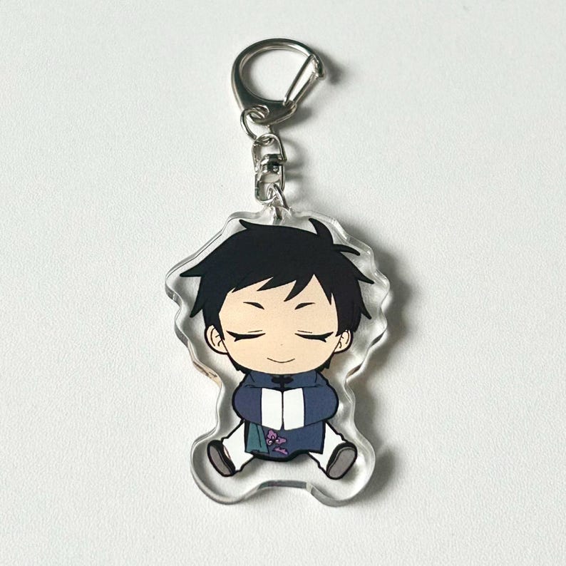 May include: A clear acrylic keychain featuring a cartoon character with black hair, wearing a blue and purple outfit. The character has closed eyes and a relaxed expression.