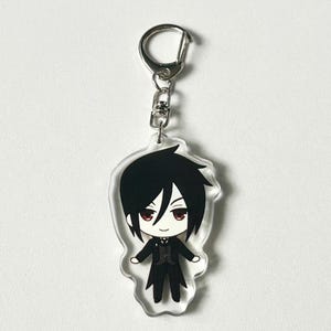 May include: A black and white anime character keychain with red eyes. The character is wearing a black suit and a white shirt with a black bow tie. The keychain is made of clear acrylic and has a silver metal ring.