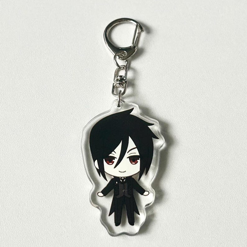 May include: A black and white anime character keychain with red eyes. The character is wearing a black suit and a white shirt with a black bow tie. The keychain is made of clear acrylic and has a silver metal ring.