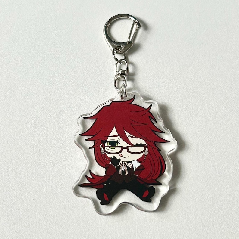May include: A red-haired anime character with green eyes and a sly smile is featured on a clear acrylic keychain. The character is wearing a black suit and a white bow tie. The keychain has a silver metal ring.