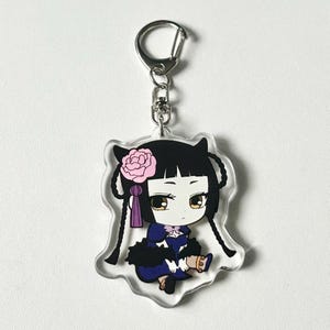 May include: A clear acrylic keychain featuring a cartoon character with black hair, a pink flower, and a purple ribbon. The character is wearing a blue and black dress and is holding a gold object.
