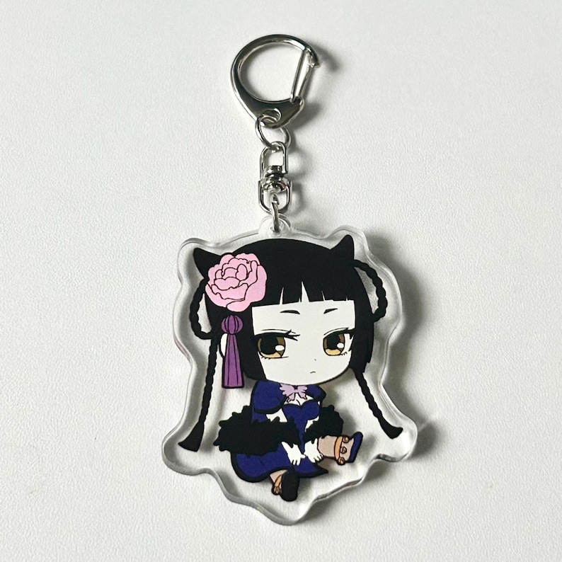 May include: A clear acrylic keychain featuring a cartoon character with black hair, a pink flower, and a purple ribbon. The character is wearing a blue and black dress and is holding a gold object.