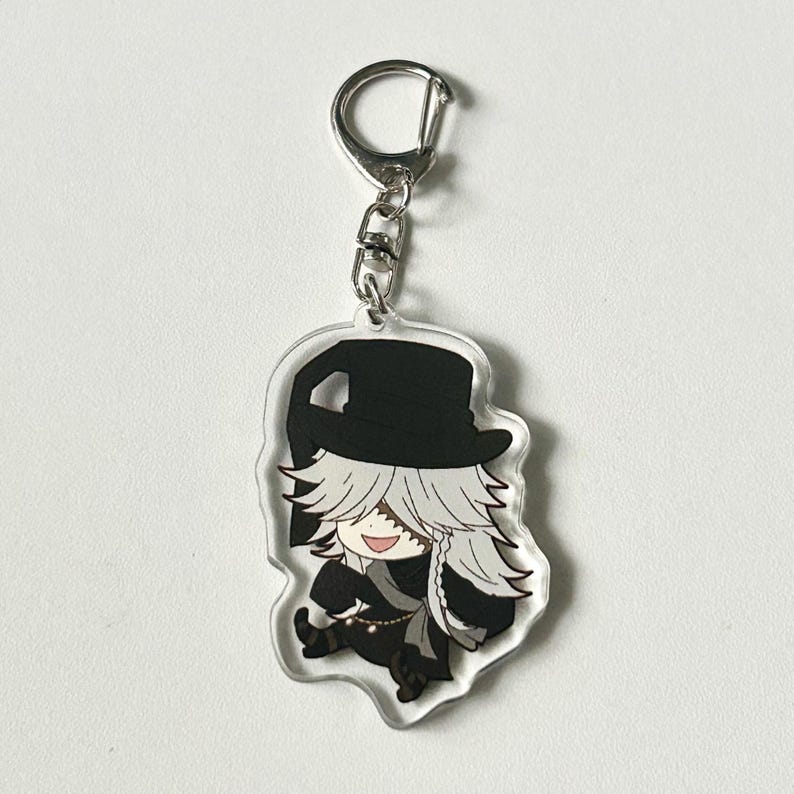 May include: A black and white anime character keychain with a white hat and long white hair. The character is smiling and wearing a black outfit. The keychain is attached to a silver metal ring.