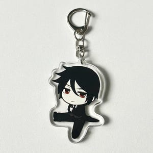 May include: A clear acrylic keychain featuring a cartoon character with black hair, a white shirt, and a black suit. The character has red eyes and a serious expression. The keychain is attached to a silver metal ring.