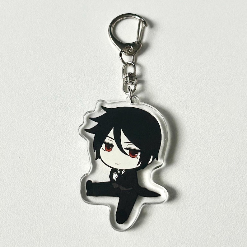 May include: A clear acrylic keychain featuring a cartoon character with black hair, a white shirt, and a black suit. The character has red eyes and a serious expression. The keychain is attached to a silver metal ring.