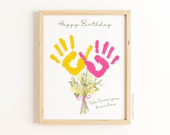 May include: A framed print with a light wood frame. The artwork features yellow and pink handprints forming a bouquet over a floral arrangement. The text "Happy Birthday" and "We love you bunches" are written on the print.
