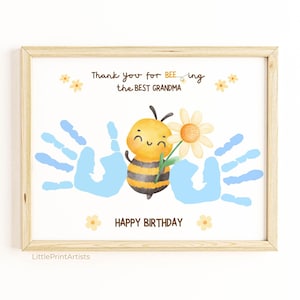 Può includere: Stampa incorniciata con un'illustrazione ad acquerello. Un'ape sorridente tiene in mano un fiore, con impronte di mani blu e il testo "Thank you for BEE-ing the best Grandma" e "Happy Birthday". La stampa ha una cornice in legno chiaro.