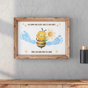 May include: A framed print featuring a watercolor illustration. The artwork depicts a smiling bee holding a flower, with blue footprints on either side. The text reads, "ICH SUMM VOR GL&Uuml;CK, WEIL ES DICH GIBT!" and "OMA, ICH HAB DICH SO LIEB!"