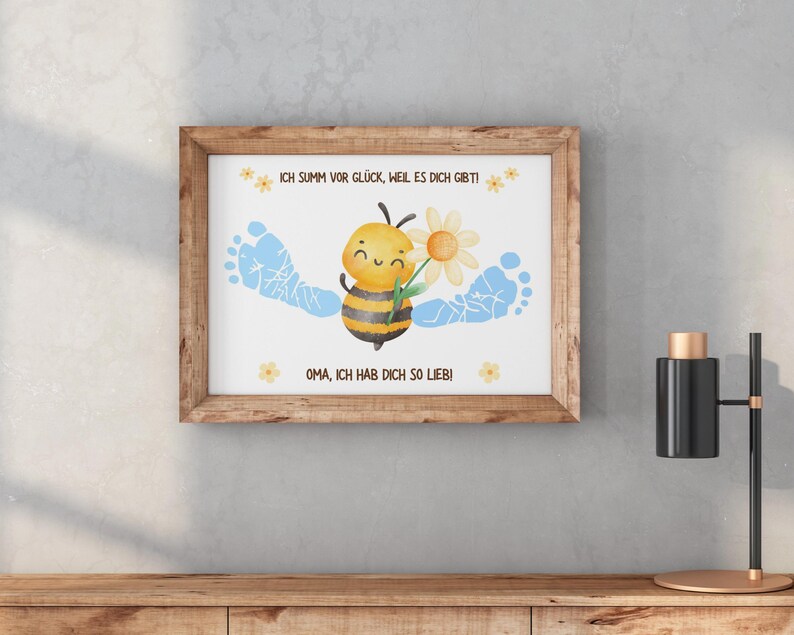 May include: A framed print featuring a watercolor illustration. The artwork depicts a smiling bee holding a flower, with blue footprints on either side. The text reads, "ICH SUMM VOR GL&Uuml;CK, WEIL ES DICH GIBT!" and "OMA, ICH HAB DICH SO LIEB!"