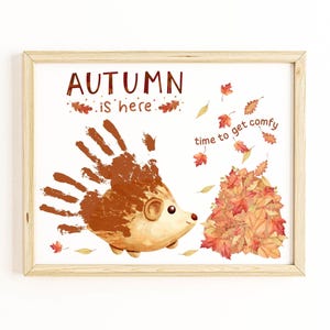 May include: A framed print featuring a handprint hedgehog illustration with the text "AUTUMN is here" and "time to get comfy." The hedgehog is brown, with a pile of autumn leaves. The frame is made of light wood.