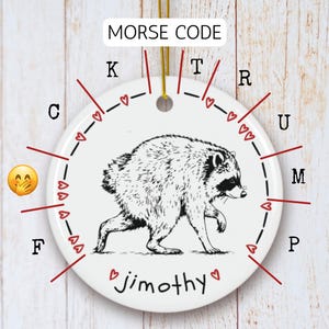 May include: A round white ceramic ornament featuring a black and white illustration of a raccoon in the center. The text MORSE CODE appears at the top, with letters and red heart icons arranged around the perimeter. The name jimothy is printed in black at the bottom, accompanied by a yellow emoji with a hand over its mouth.