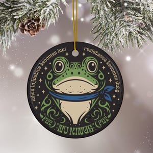 May include: A round black ceramic ornament featuring a green frog illustration wearing a blue neckerchief. The text reads: When in justice becomes law resistance becomes duty, and NO KINGS. It includes a gold hanging string and a small hole at the top.