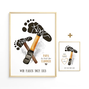 May include: Two framed prints with a light wood frame. The larger print features two crossed hammers with a footprint design and the text "PAPA, du bist der HAMMER" and "WIR HABEN DICH LIEB". The smaller print has a single hammer and footprint with text.