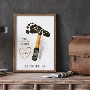 May include: Framed art print with a hammer design made from a footprint. The print includes the text "Papa du bist der Hammer" and "Ich hab dich lieb", with a heart and the name "Elias". The frame is brown.