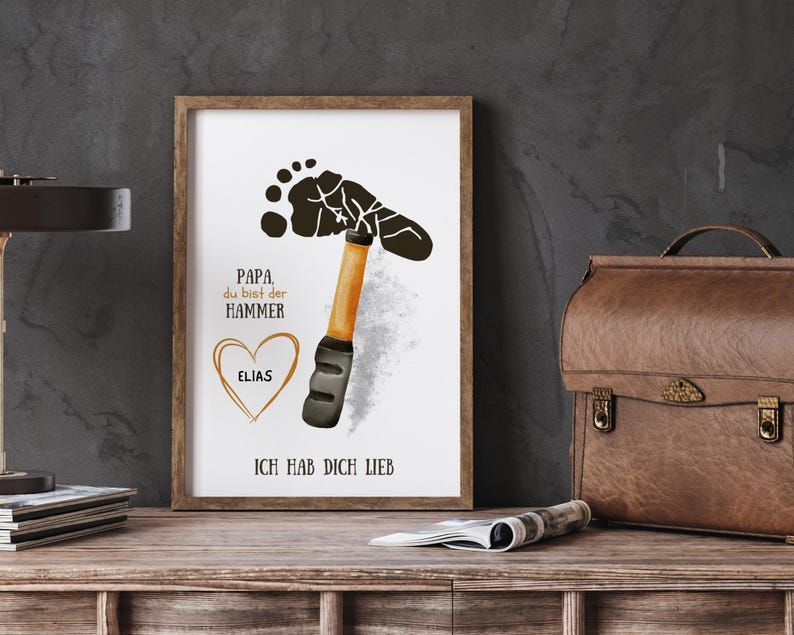 May include: Framed art print with a hammer design made from a footprint. The print includes the text "Papa du bist der Hammer" and "Ich hab dich lieb", with a heart and the name "Elias". The frame is brown.