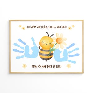 May include: A framed print featuring a watercolor illustration. A smiling bee holds a daisy between two blue handprints. The text reads "ICH SUMM VOR GLÜCK, WEIL ES DICH GIBT!" and "OMA, ICH HAB DICH SO LIEB!" in German. The frame is light brown.