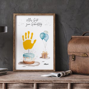 Pu&ograve; includere: Una stampa ad acquerello incorniciata con un tema di compleanno. L'opera presenta un'impronta di mano gialla, una torta, palloncini blu e un regalo incartato. Il testo "Alles Gute zum Geburtstag" &egrave; scritto in alto, con il nome "Maxi" in basso.