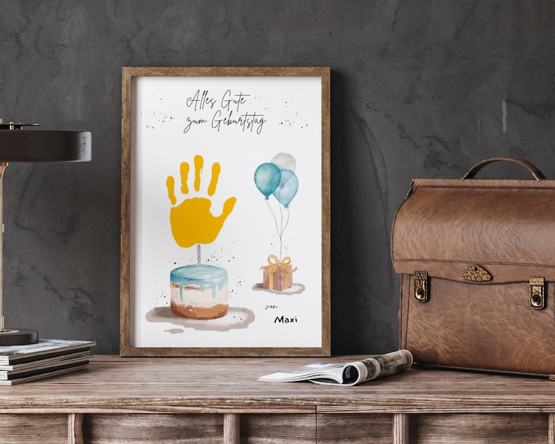 Pu&ograve; includere: Una stampa ad acquerello incorniciata con un tema di compleanno. L'opera presenta un'impronta di mano gialla, una torta, palloncini blu e un regalo incartato. Il testo "Alles Gute zum Geburtstag" &egrave; scritto in alto, con il nome "Maxi" in basso.