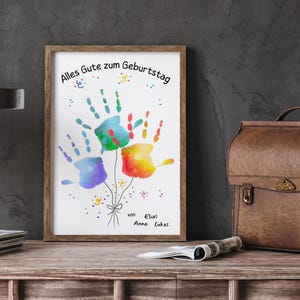 Peut inclure: Une impression encadr&eacute;e avec des empreintes de mains color&eacute;es en forme de ballons. L'&oelig;uvre comprend le texte allemand "Alles Gute zum Geburtstag" et les noms "Elias, Anna, Lukas". Les empreintes sont bleues, vertes, rouges et jaunes.